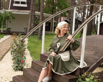 May include: A woman wearing an olive green dress with a white lace apron and headscarf sits on wooden steps. The dress has long sleeves and a white lace trim. She wears gray shoes. A windmill is in the background.