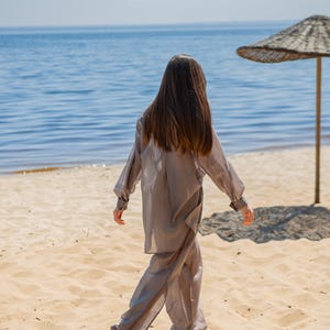 May include: A person wearing a light gray, flowing two-piece outfit walks on a sandy beach towards the ocean. The outfit includes a long-sleeved top and wide-leg pants. A straw umbrella provides shade on the beach.