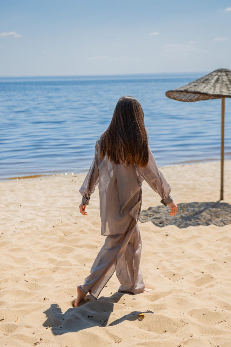 May include: A person wearing a light gray, flowing two-piece outfit walks on a sandy beach towards the ocean. The outfit includes a long-sleeved top and wide-leg pants. A straw umbrella provides shade on the beach.