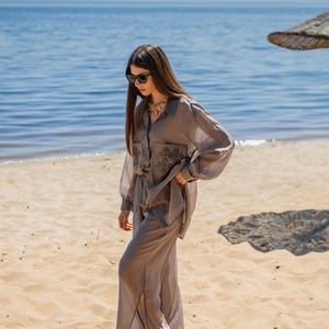 May include: A light gray, long-sleeved shirt and matching wide-leg pants set. The shirt has a button-down front and embroidered details on the pockets. The outfit is worn on a sandy beach with the ocean in the background.