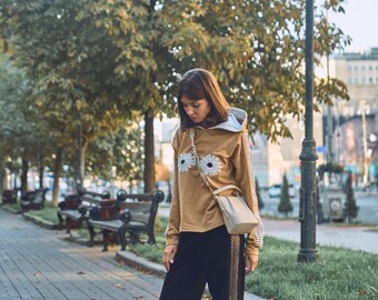 May include: A woman wearing a brown hoodie with white flower embroidery, black pants with a gold stripe, and a white handbag.