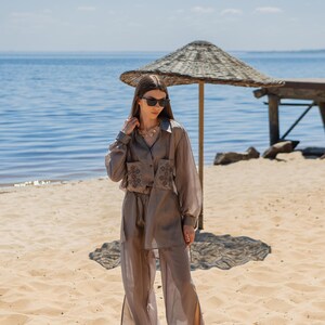 May include: A light brown, two-piece outfit on a sandy beach. The outfit includes a long-sleeved button-down shirt with embroidered details and matching wide-leg pants. The model is wearing sunglasses and a necklace. The background features a beach umbrella and the ocean.