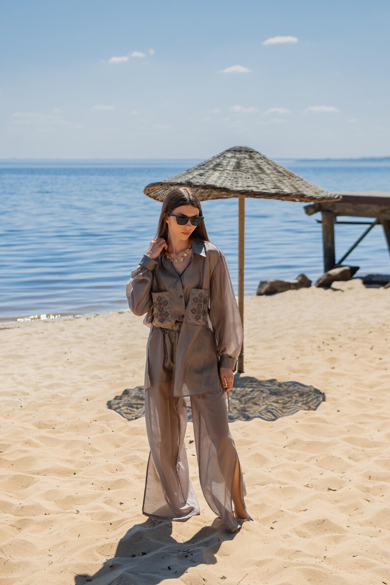 May include: A light brown, two-piece outfit on a sandy beach. The outfit includes a long-sleeved button-down shirt with embroidered details and matching wide-leg pants. The model is wearing sunglasses and a necklace. The background features a beach umbrella and the ocean.