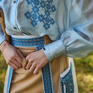 K&ouml;nnte beinhalten: Hellblaues Hemd mit gestickten blauen geometrischen Mustern auf der Tasche. Das Outfit umfasst einen beigefarbenen Rock mit blau-wei&szlig;em Musterbesatz und einen goldenen Sonnenring.
