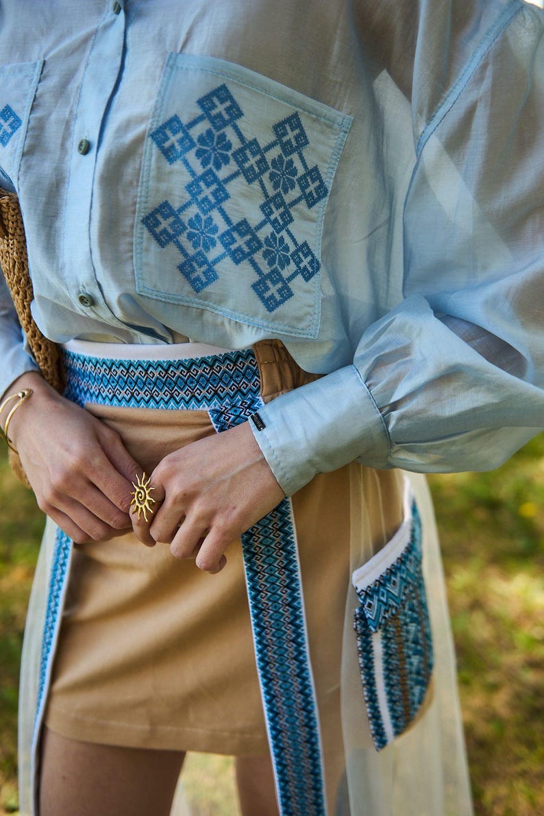 K&ouml;nnte beinhalten: Hellblaues Hemd mit gestickten blauen geometrischen Mustern auf der Tasche. Das Outfit umfasst einen beigefarbenen Rock mit blau-wei&szlig;em Musterbesatz und einen goldenen Sonnenring.