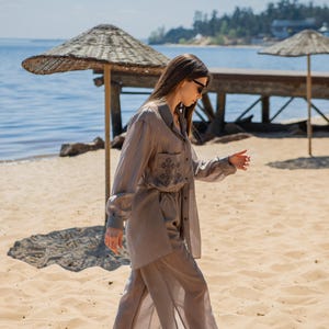 May include: A light brown linen two-piece set, including a button-down shirt with a pocket and embroidered detail, and wide-leg pants. The outfit is worn on a sandy beach near the water, with a straw umbrella and wooden pier in the background.