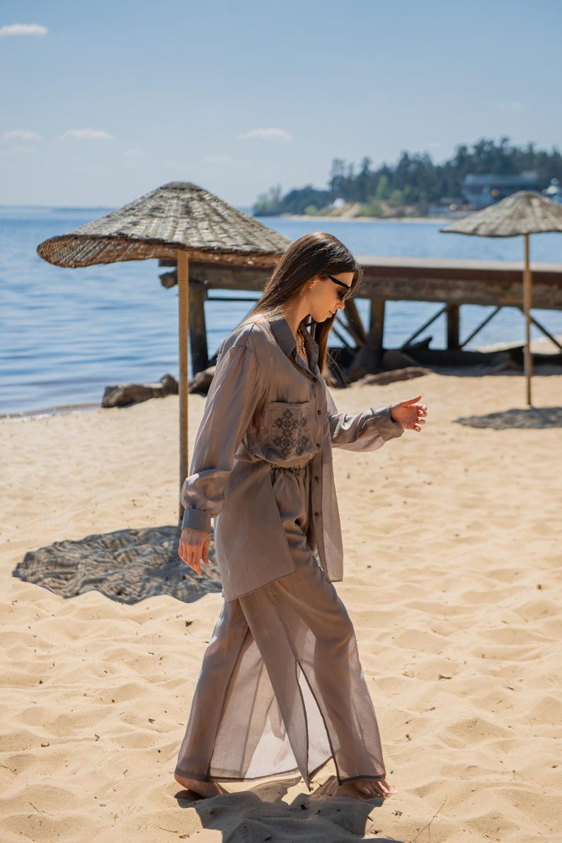 May include: A light brown linen two-piece set, including a button-down shirt with a pocket and embroidered detail, and wide-leg pants. The outfit is worn on a sandy beach near the water, with a straw umbrella and wooden pier in the background.