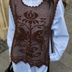 May include: A brown, open-weave crochet vest with a floral design. The vest is worn over a light blue button-down shirt. The vest has a scalloped edge and a detailed pattern. The overall style is bohemian and handmade.