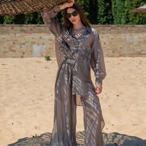 May include: A light gray, semi-sheer button-down shirt and wide-leg pants set. The shirt has embroidered detailing and is paired with matching pants. The outfit is worn on a sandy beach, with a woven sunshade overhead.