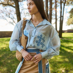 K&ouml;nnte beinhalten: Hellblaues, durchsichtiges Hemd mit Kn&ouml;pfen und gestickten Details auf den Taschen. Das Outfit umfasst einen beigefarbenen Rock mit blauem, gesticktem Besatz, eine Strohtasche und Goldschmuck. Das Model tr&auml;gt einen goldenen Ring in Sonnenform.