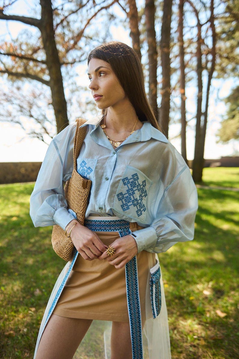 K&ouml;nnte beinhalten: Hellblaues, durchsichtiges Hemd mit Kn&ouml;pfen und gestickten Details auf den Taschen. Das Outfit umfasst einen beigefarbenen Rock mit blauem, gesticktem Besatz, eine Strohtasche und Goldschmuck. Das Model tr&auml;gt einen goldenen Ring in Sonnenform.
