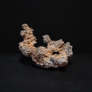 Op de afbeelding: Een grote, onregelmatig gevormde rots met een ruw, getextureerd oppervlak. De rots is lichtbruin van kleur met hints van grijs en heeft een gat in het midden.