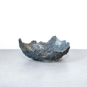 May include: A handcrafted, dark gray bowl with a textured, organic shape. The bowl has a rough, uneven surface and a unique, sculptural design. The interior appears to have a lighter, metallic finish. The bowl sits on a light gray surface against a white background.
