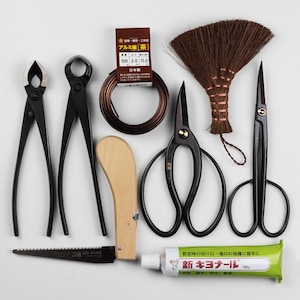 Puede incluir: Un conjunto de herramientas de bonsái que incluye tijeras de poda, una sierra, una herramienta de madera, una bobina de alambre, un cepillo y un tubo de sellador para árboles. El tubo de sellador está etiquetado como "新キヨナール 100g".