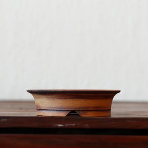 Peut inclure: Un pot à bonsaï en céramique brune de forme arrondie avec un bord légèrement évasé. Le pot a une surface texturée et une petite ouverture au fond pour le drainage.