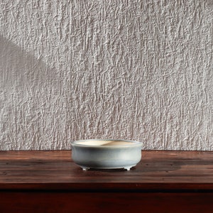Peut inclure: Petit pot à bonsaï rond en céramique bleu clair, avec trois petits pieds. L'intérieur est blanc crème. Le pot est posé sur une surface en bois brun foncé, devant un mur blanc texturé. Idéal pour les petites plantes.