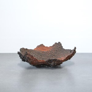 May include: A textured, handmade ceramic bowl with a unique, organic shape. The bowl features a dark brown exterior with reddish-brown and black accents. The interior has a reddish-brown hue. The bowl sits on a gray surface against a white background.