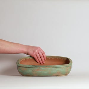 May include: A rectangular, weathered, green ceramic planter with rounded corners. The interior is a reddish-brown color. The planter is empty and ready for planting. The image shows a hand reaching into the planter.