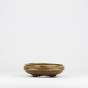 Może przedstawiać: Mała, okrągła, brązowa ceramiczna doniczka do bonsai z szeroką, płytką misą i trzema małymi nóżkami. Doniczka ma teksturowaną powierzchnię i prosty design, odpowiedni dla miniaturowych drzew i roślin.