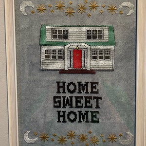 May include: A framed cross-stitch design featuring a white house with a red door and green roof. The house is surrounded by stars and crescent moons. The text "HOME SWEET HOME" is below the house.