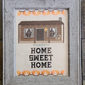 May include: A framed cross-stitch with a brown cabin and the text "Home Sweet Home" in black. The cabin has a brown roof, two windows, and a door. There are orange leaves on vines above and below the cabin.