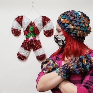 May include: A person wearing a colorful crocheted hat and matching fingerless gloves. The hat and gloves have a scale-like pattern. The person is wearing a pink and black plaid shirt and a black face mask.