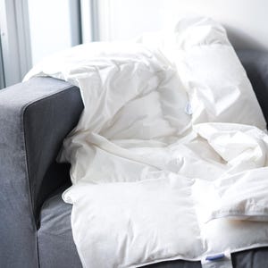May include: A white down comforter is folded and placed on a grey sofa. The comforter has a white tag with blue writing on it.
