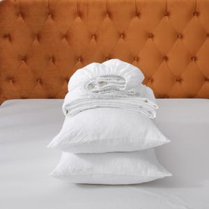 May include: White bed linen set, including a fitted sheet, flat sheet, and two pillowcases, neatly arranged on a white bed. An orange-brown, tufted headboard is in the background. The bedding is folded.