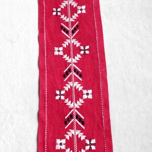 Könnte beinhalten: Ein roter Tischläufer aus Stoff mit weißen und schwarzen Stickereien. Das Design zeigt ein sich wiederholendes Muster aus geometrischen Formen und floralen Motiven.