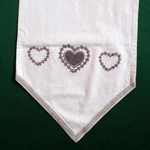 Op de afbeelding: Een witte tafelrunner met een bruin geborduurd ontwerp van drie harten. De harten zijn omrand met een decoratieve steek.