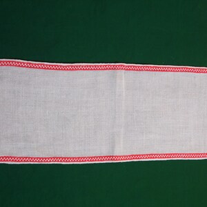 May include: White linen table runner with a red embroidered border. The border features a repeating pattern of stylized figures.