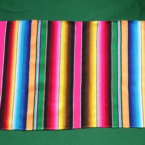 Puede incluir: Una colorida manta mexicana con rayas de morado, rosa, naranja, amarillo, verde, azul y negro. La manta tiene flecos blancos en los extremos.
