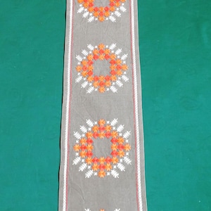 May include: A grey table runner with a white and orange embroidered design. The design features five circular patterns of flowers.