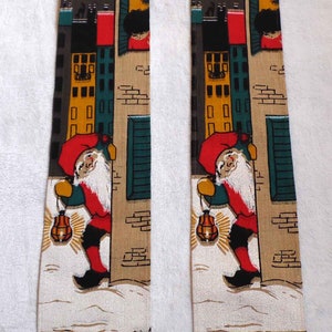 May include: Two vintage wall hangings featuring a whimsical scene of a gnome holding a lantern in front of a brick building. The gnome is wearing a red hat, red jacket, and black pants. The building is decorated with yellow and green windows. The background is a beige and brown fabric.