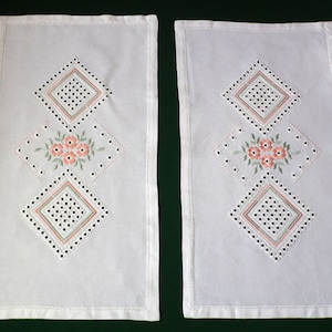 Puede incluir: Dos caminos de mesa bordados blancos con un patrón de diamante y flores rosas. Los caminos de mesa están hechos de una tela delicada y tienen un borde festoneado.