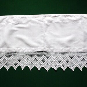 Op de afbeelding: Witte valance met een decoratieve, opengewerkte kanten rand. De stof is effen wit en het kant heeft een herhalend ruitpatroon met een geschulpte rand. De valance is waarschijnlijk voor een raambekleding.