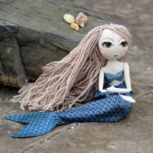 Könnte beinhalten: Eine handgefertigte Meerjungfrauenpuppe mit langen, hellbraunen Garnhaaren, einem blau-grünen gemusterten Schwanz und einem blauen Stoffoberteil. Die Puppe sitzt auf einem Felsen mit Muscheln in der Nähe.