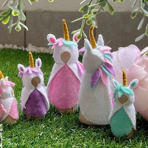 Puede incluir: Cinco muñecas de fieltro de unicornio con cuernos dorados y capas de diferentes colores. Las muñecas están dispuestas sobre una superficie verde con una flor rosa en el fondo.