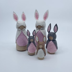 May include: A set of five felt bunny finger puppets. The puppets are all different sizes and have pink, white and grey felt. The puppets are wearing pink felt capes.