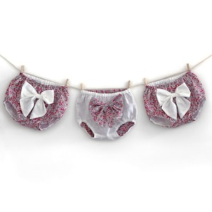 Pode incluir: Três pares de bloomers de algodão branco com estampa floral rosa e branca e um laço branco na frente. Os bloomers estão pendurados em um varal.