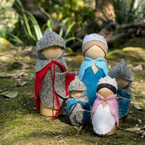 May include: Five wooden peg dolls dressed in felt clothing. The dolls are wearing a variety of colours, including grey, red, blue, pink, and white. The dolls are standing on a bed of moss.