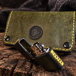 Könnte beinhalten: Ein olivgrünes Lederetui für Feuerzeuge mit gelben Nähten und zwei schwarzen Metalldruckknöpfen, versehen mit einem runden Logo, das ein Vogelprofil und den Text THE FLOCK HANDMADE zeigt. Ein schwarzes Metallfeuerzeug liegt vor dem Etui.