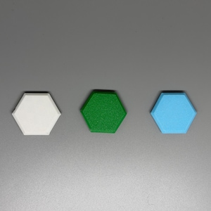 May include: Three hexagonal objects in white, green, and light blue are arranged horizontally against a gray background. The objects appear to be made of a textured material, with the green object having the most visible texture.