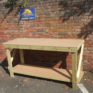 May include: A wooden workbench with a light brown tabletop and a lower shelf. The frame is made of light green wood. The workbench is set against a red brick wall. The sign on the wall reads "Wells Timber Products Parking Only."
