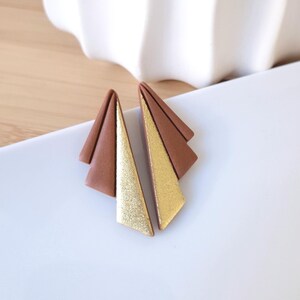 May include: A pair of geometric earrings. Each earring features a brown, triangular shape with a gold, glittery triangle attached. The earrings are set against a white background, with a glimpse of a white, textured vase in the background.