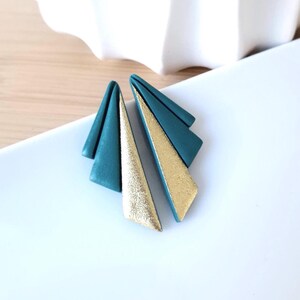 May include: A pair of teal and gold geometric earrings. Each earring features a teal triangle with a gold metallic triangle overlay. The earrings are displayed on a white surface, showcasing their modern design and color contrast.