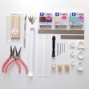 May include: A collection of polymer clay tools and supplies, including pink-handled pliers, clay cutters, FIMO clay in brown, beige, and teal, a drill, and various other crafting tools. The tools are arranged on a white surface.