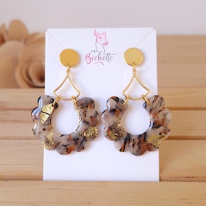 Pode incluir: Um par de brincos com um design floral. Os brincos apresentam um pino dourado e uma forma geométrica dourada que se conecta a um pingente em forma de flor com um padrão marmorizado em tons de marrom, preto e dourado. A marca "ma Bichette" é visível.