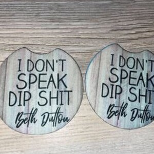 May include: Two round wooden coasters with a light wood grain pattern. The coasters have the text "I don't speak dip shit" and the name "Beth Dutton" in black lettering.
