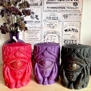 May include: Three pillar candles in red, purple, and black, each with a detailed eye design and hands. The candles have a textured surface and are part of a witchy or mystical theme.